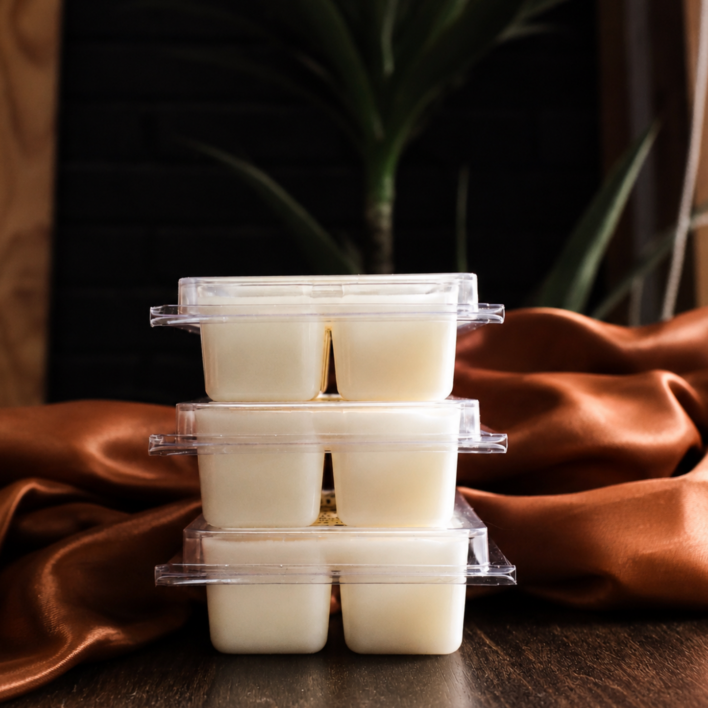Three stacked clear plastic clamshell containers holding pale creamy white cubes, arranged on draped copper-brown satin fabric over a dark wooden surface with a blurred plant in the background.