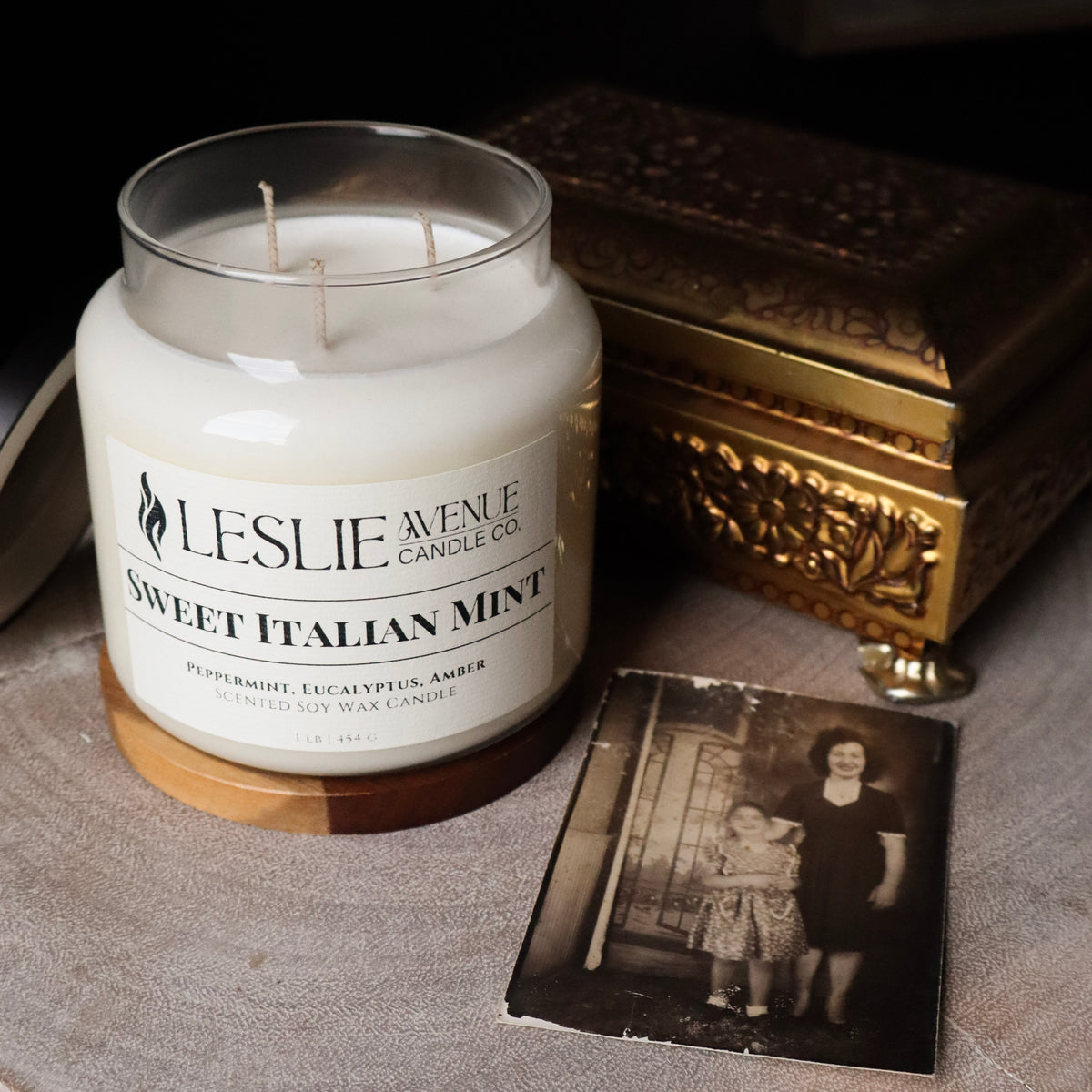 A jar candle labeled 'Leslie Avenue Candle Co. Sweet Italian Mint' sits on a tabletop beside an ornate gold-toned box and a vintage black-and-white photo card.