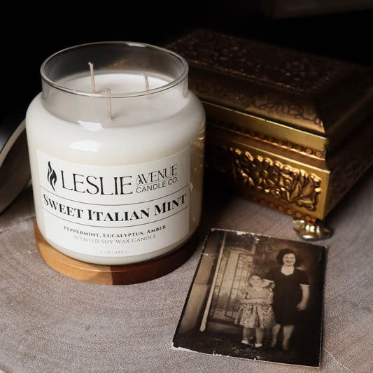 A jar candle labeled 'Leslie Avenue Candle Co. Sweet Italian Mint' sits on a tabletop beside an ornate gold-toned box and a vintage black-and-white photo card.