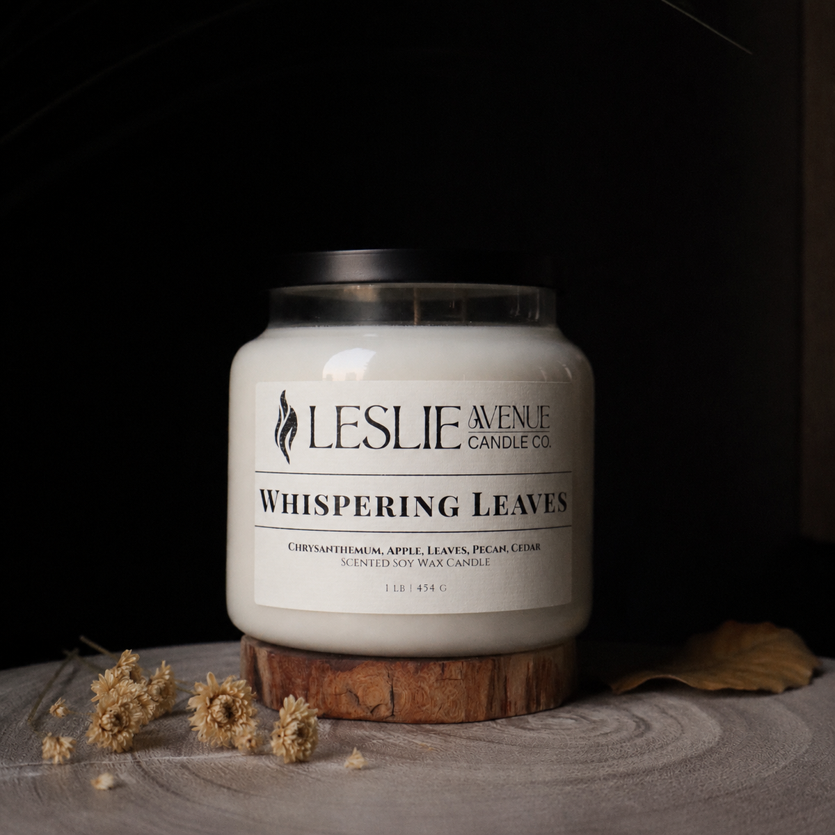 A white wax candle in a clear glass jar with a black lid, displayed on a rustic wood slice coaster. The label reads 'Leslie Avenue Candle Co' and 'Whispering Leaves'.