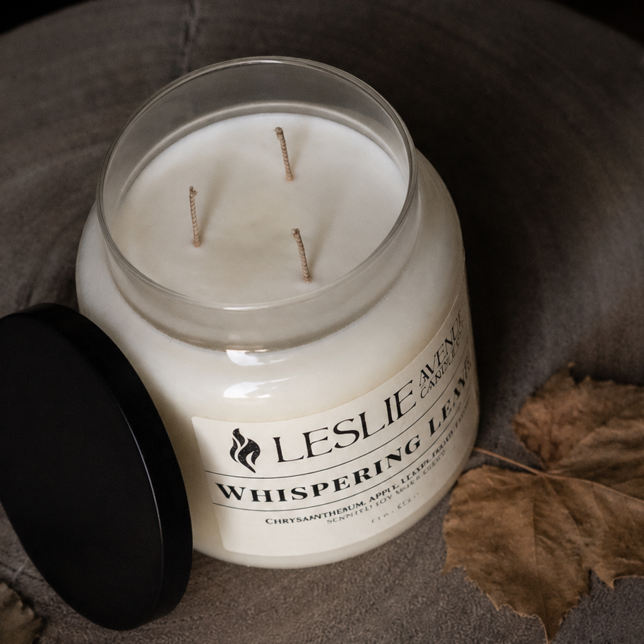A three-wick soy candle in a clear glass jar with white wax and a black lid resting beside it on a textured gray surface, surrounded by dried leaves. The label reads 'Leslie Rose Whispering Pine'.