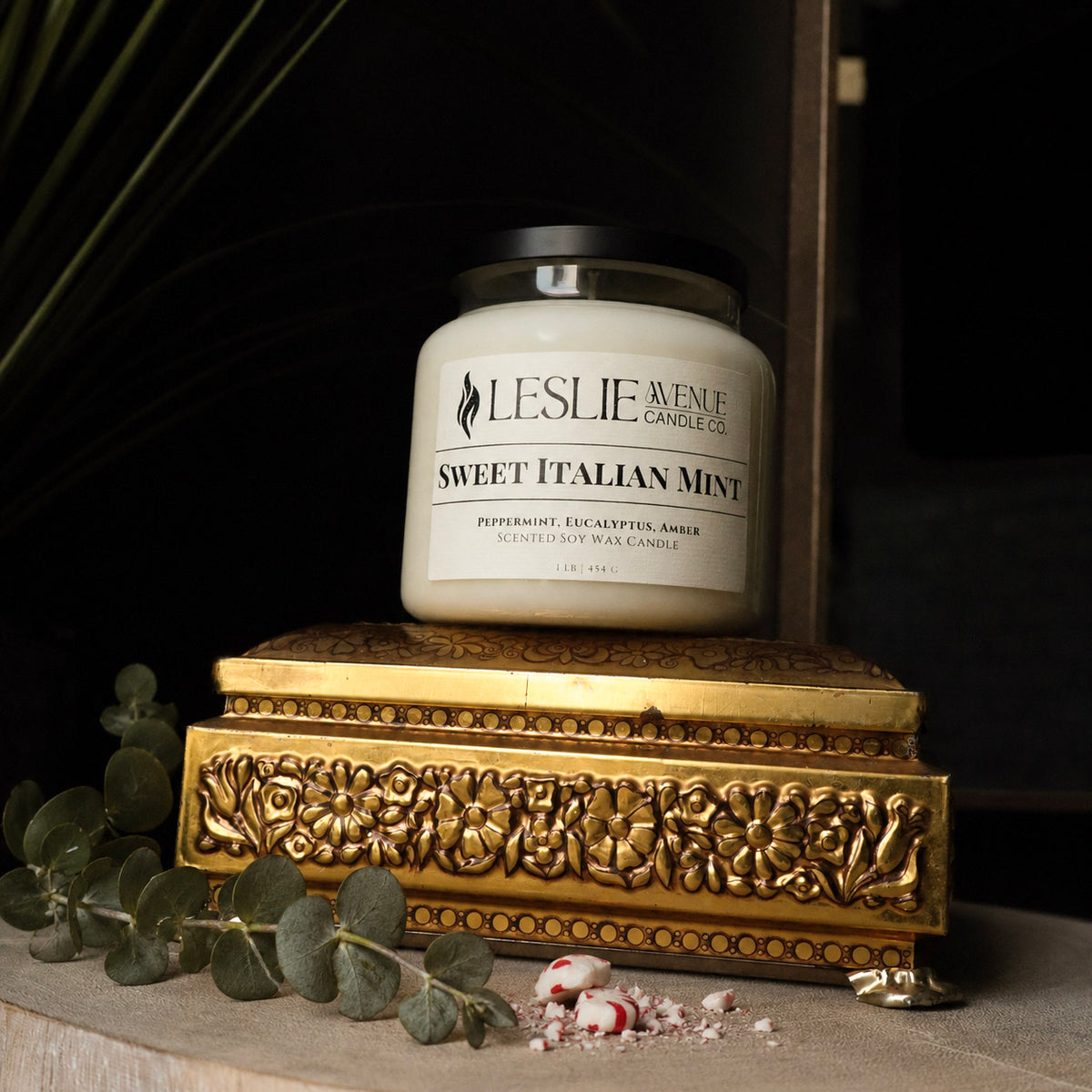 A jar candle labeled 'Leslie Avenue Candle Co. Sweet Italian Mint' sits on an ornate gold decorative box with eucalyptus leaves and candy pieces around it in a dark styled setting.
